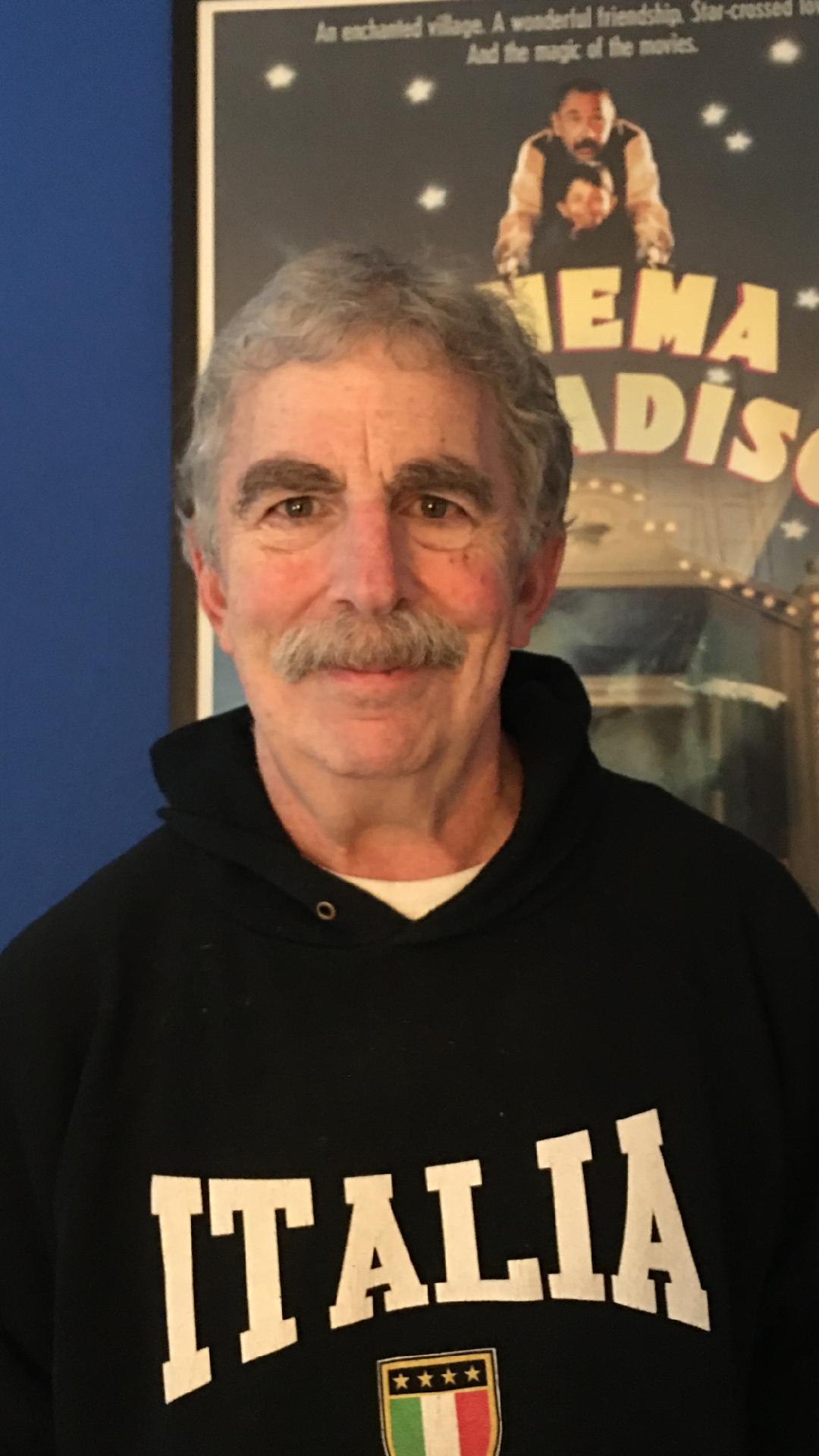 older white man with bushy eyebrows and a mustache wearing a black hoodie that says Italia standing in front of a movie poster