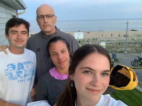 A white family of four the man is bald with glasses wearing a gray hoodie and holding a yellow hat