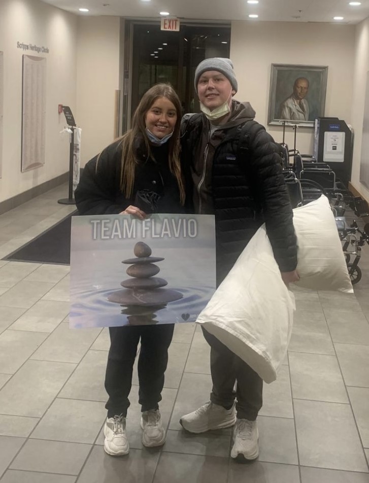 Flavio young hispanic couple she is holding Team Flavio poster and he is holding a pillow both wearing black jackets