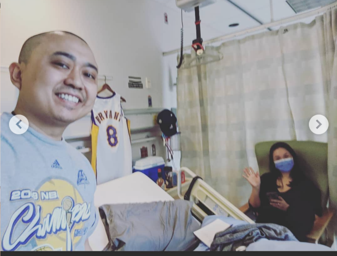 Nino Asian man in hospital room wearing blue tshirt standing by a Kobe Bryant jersey with his wife sitting in a chair waving and wearing a blue mask and dark shirt