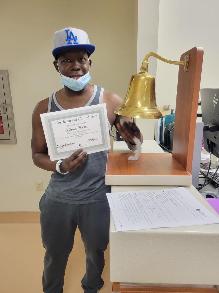 middle aged black man wearing LA ballcap and gray and white tank top and gray sweat pants holding a certificate and ringing a bell.