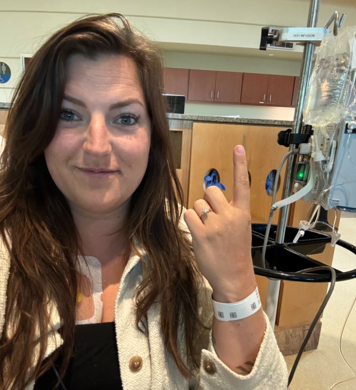 young white woman with long brown hair wearing a black shirt and white jacket sitting in a hospital room holding up her pointer finger