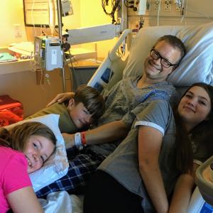 middle aged white man wearing glasses and a hospital gown sitting on a hospital bed with his son and two daughers