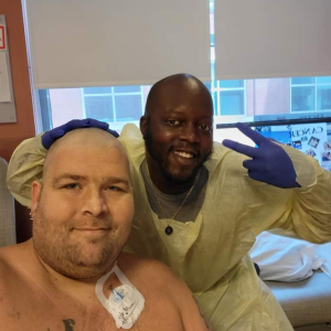 younger white male with shower cap on scruffy beard and no shirt sitting in front of black male medical tech giving a peace sign