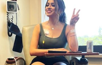 young hispanic woman sitting in a chair giving the peace sign, hair in ponytail, gray tank and black shorts, red and white tennis shoes