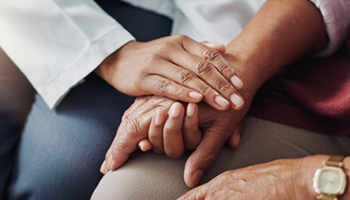 A person with dark-skinned hands clasps the hand of a second dark-skinned person to offer comfort.