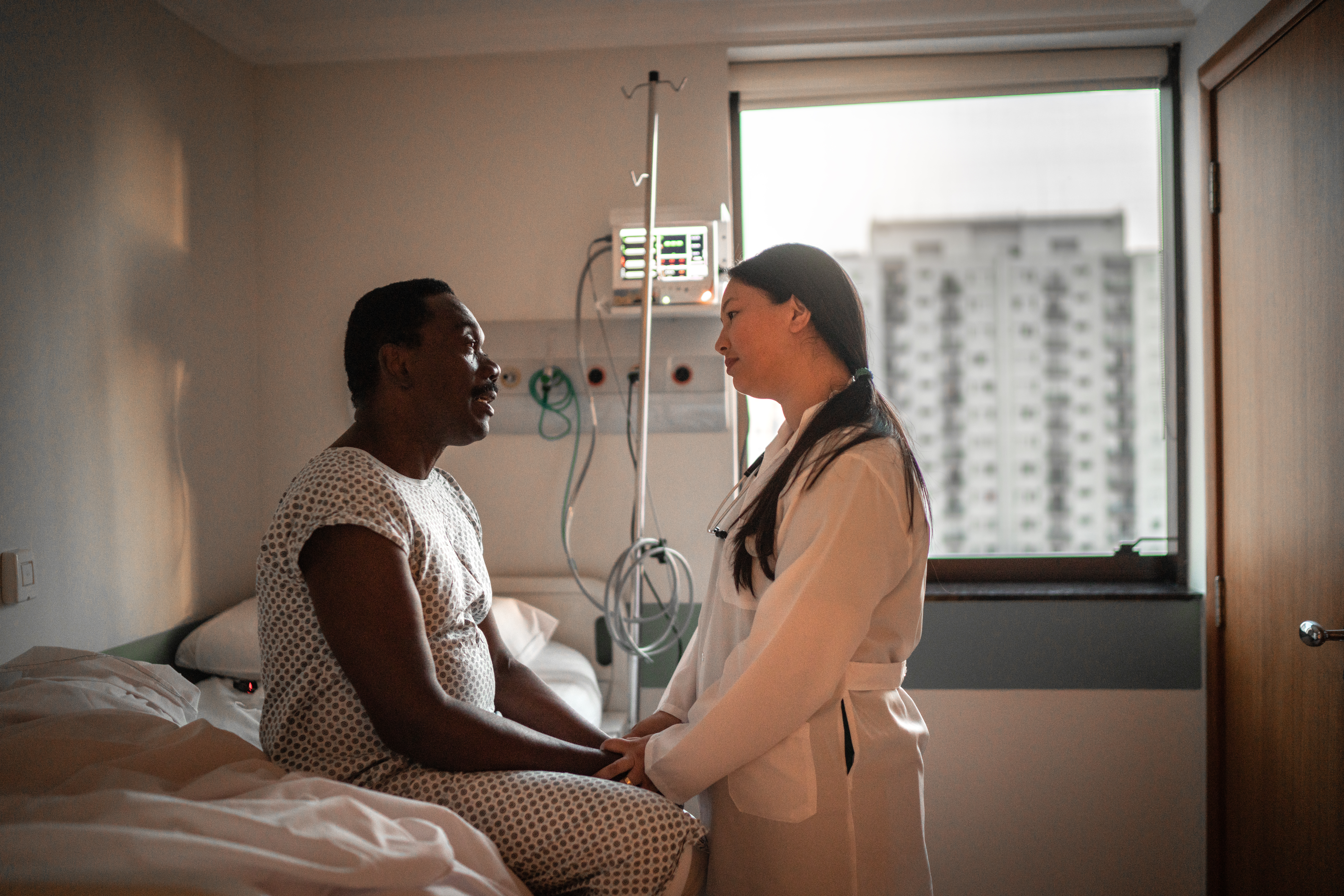 An African-American patient holds the hand and speaks with a doctor of Asian descent in a hospital room