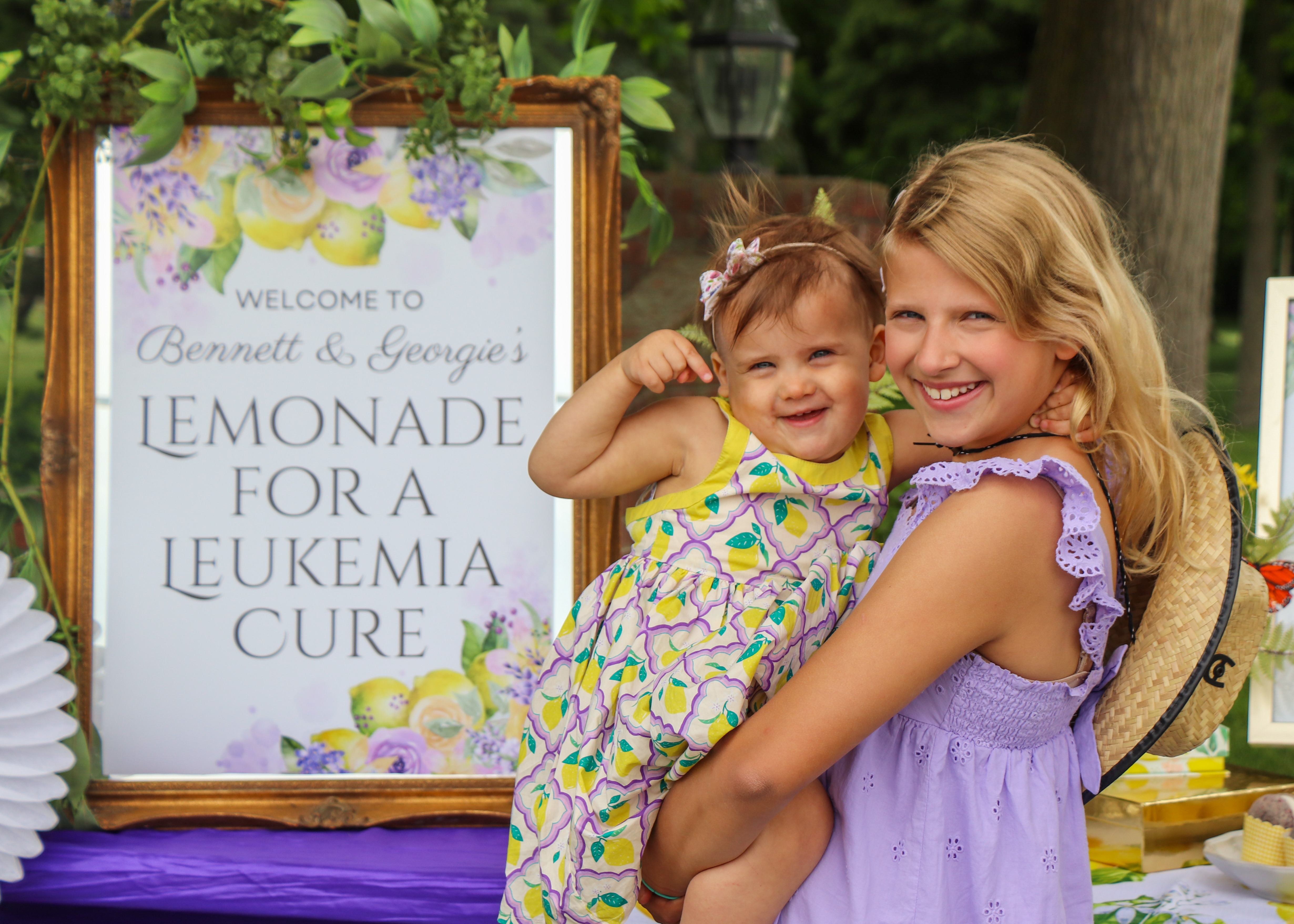 Blond girl in purple dress holding younger girl by lemonade stand sign.