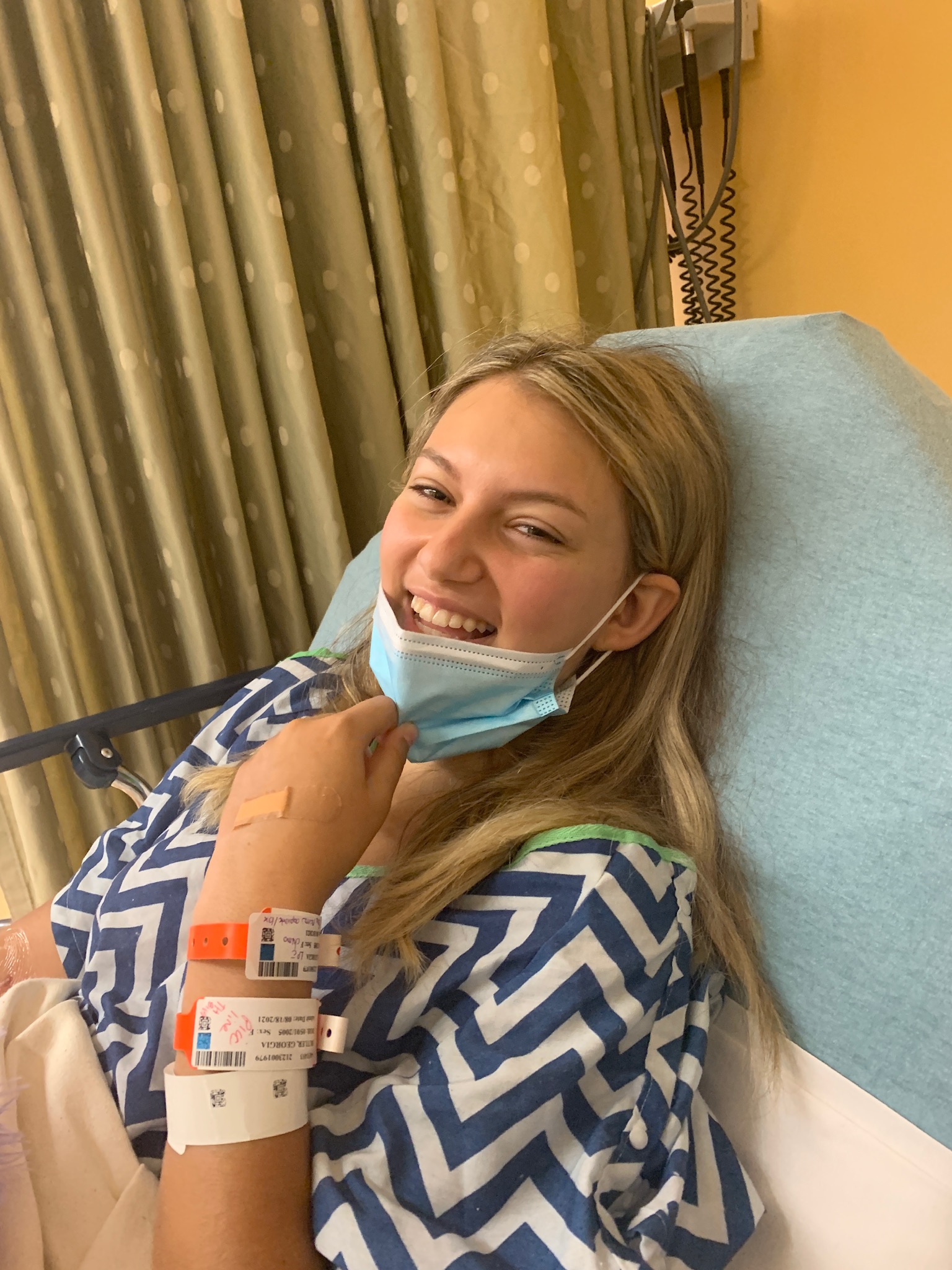 Blonde teen girl with leukemia in blue and white medical gown pulling down a face mask