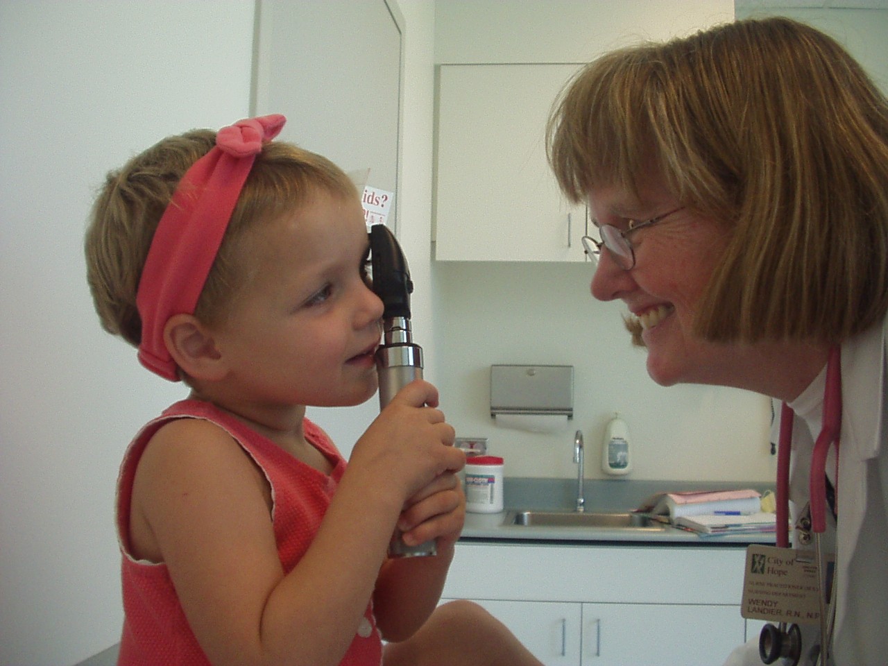 Little girl with red headband and shirt holding medical instrument next to a woman with glasses