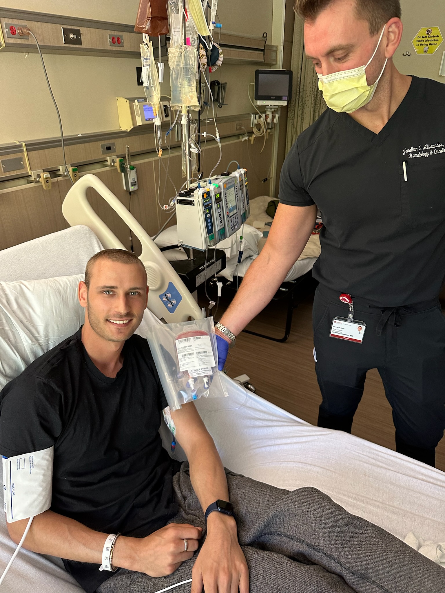 Young man in black t-shirt with leukemia in a hospital bed with a medical professional standing next to him wearing a mask