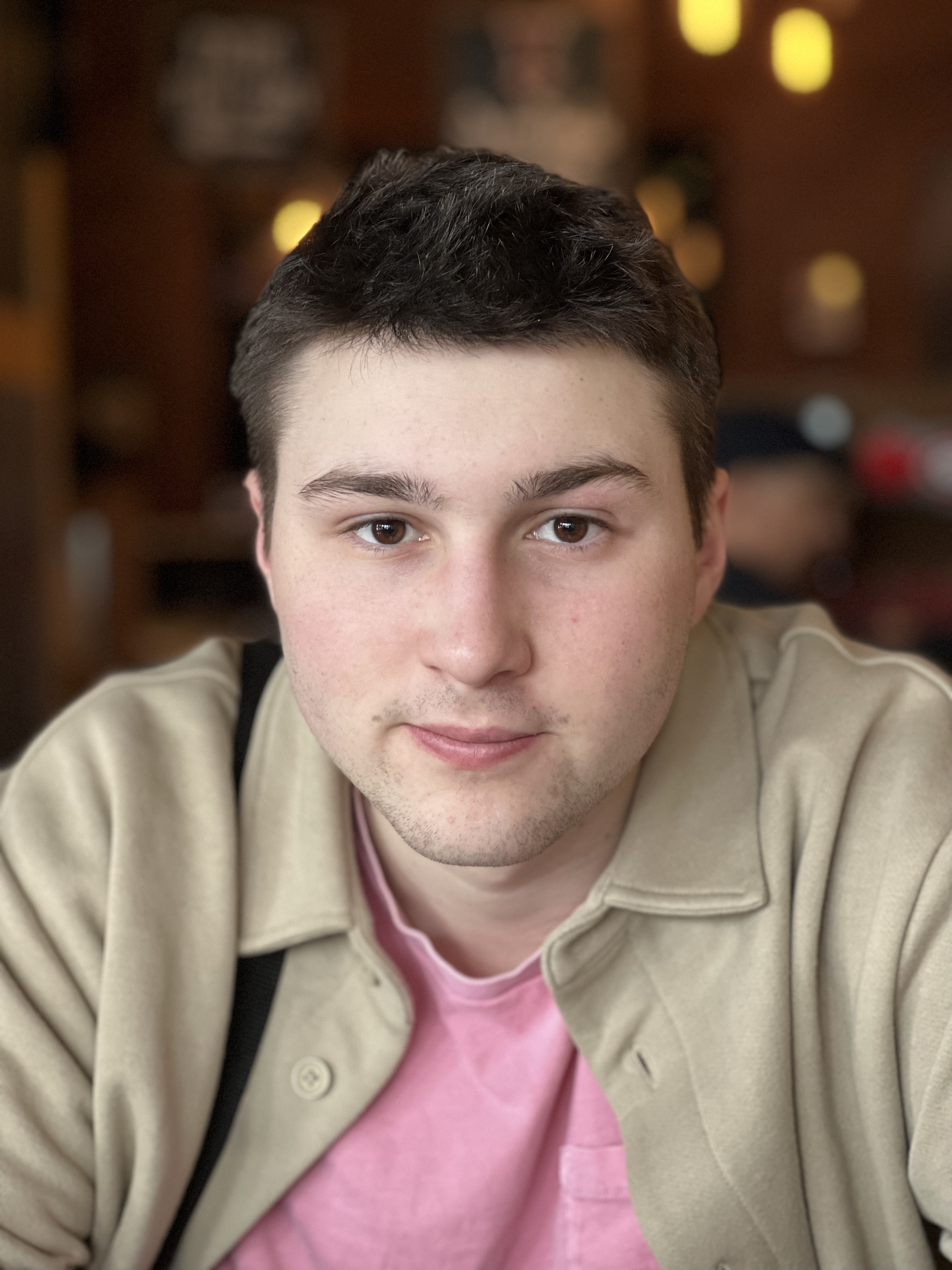 young white man with dark brown hair and chin stubble wearing a pink shirt and beige jacket