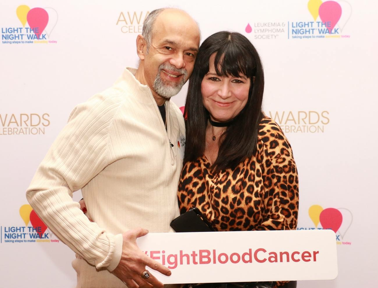 an older hispanic couple hugging in front of LTN wall he in a cream shirt holding a sign that says FightBloodCancer and her in an animal print top