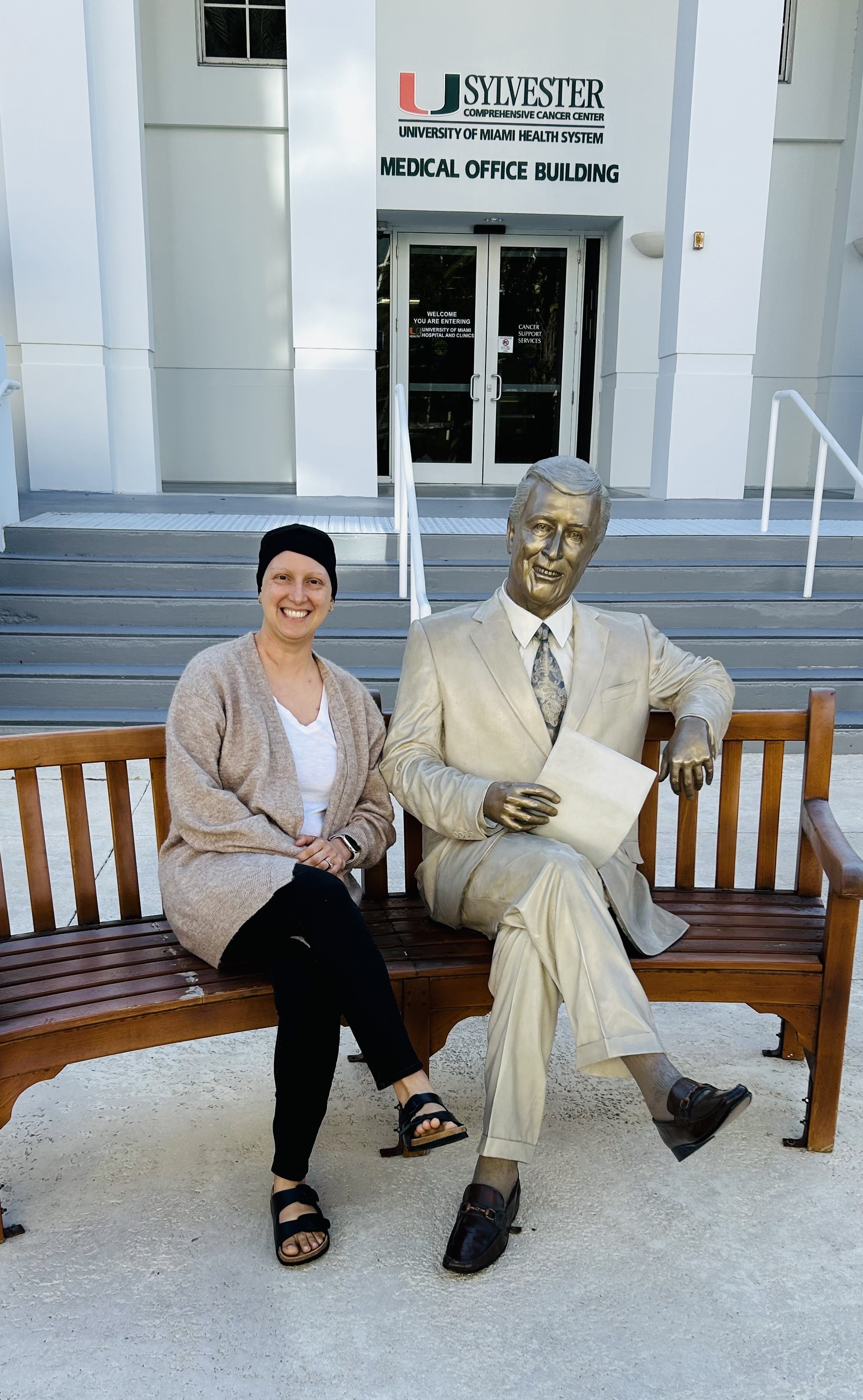 bald white woman wearing a knit cap tan sweater and white shirt sitting on a bench next to a statue