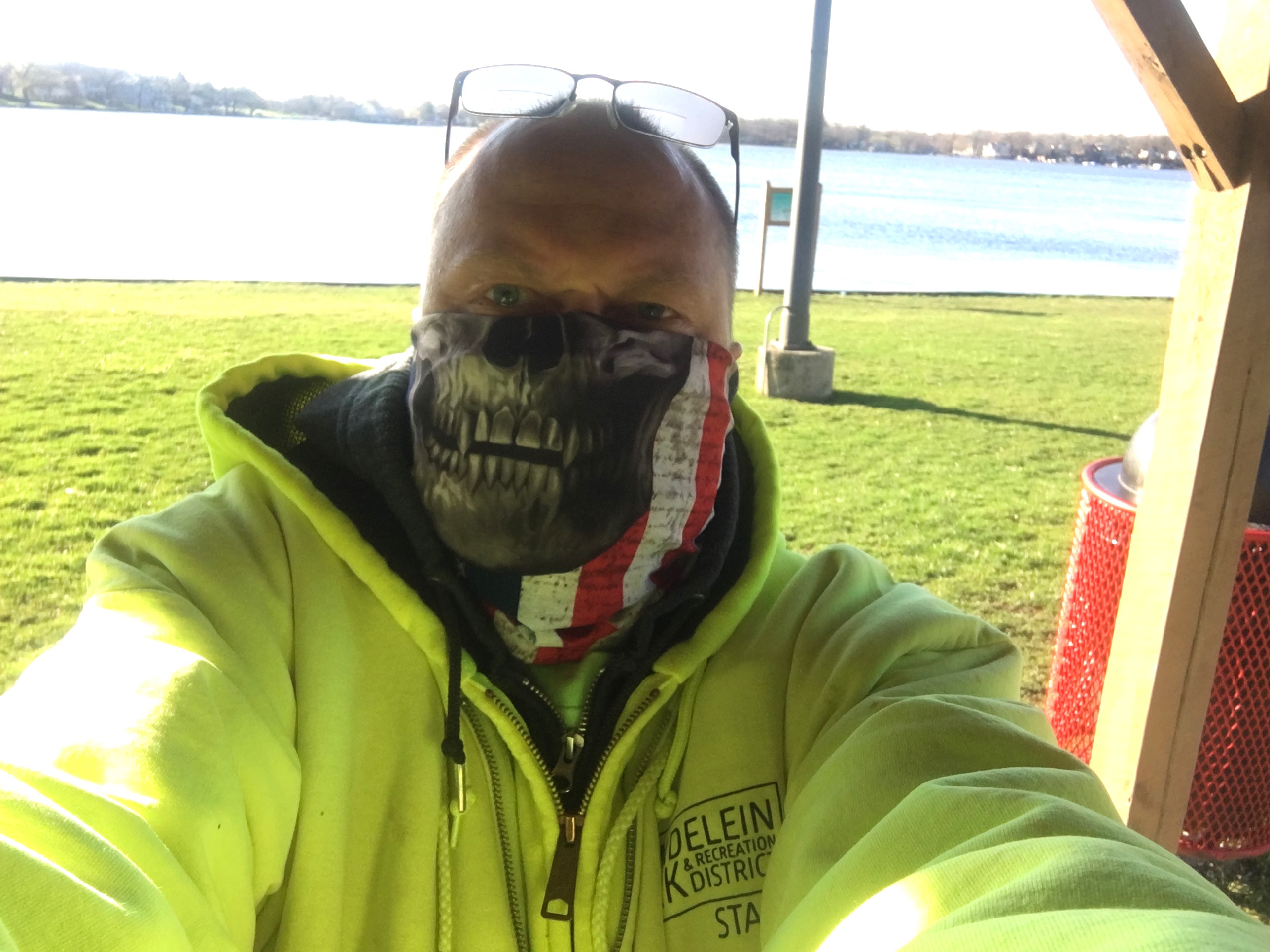 balding white man with sunglasses on his head wearing a monster mask and yellow hoodie in front of water