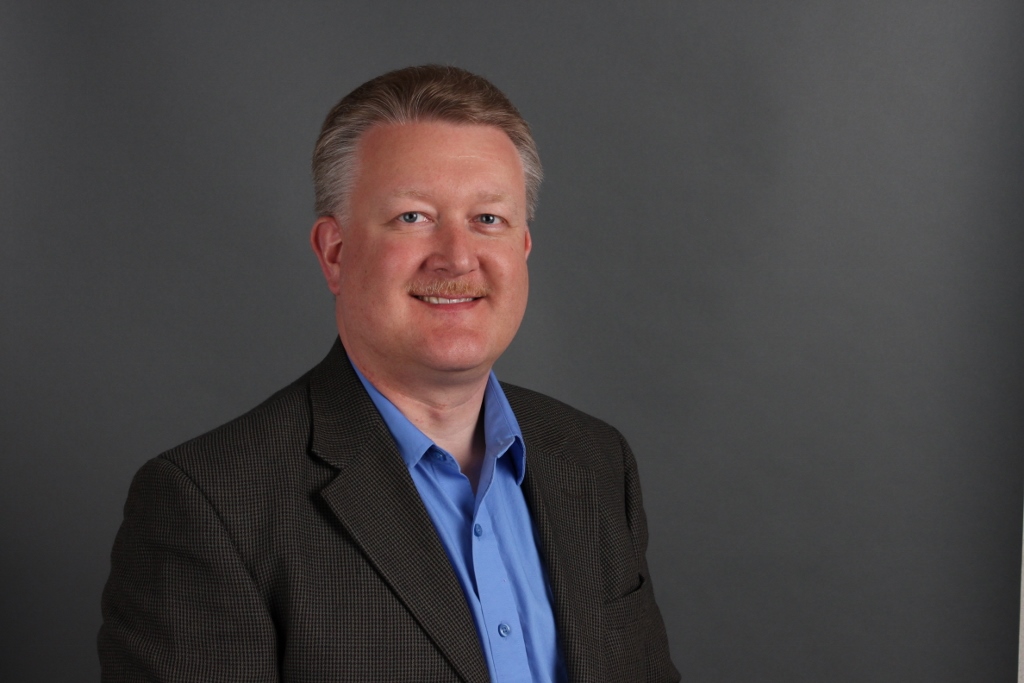 smiling middle aged white man with graying hair and mustache wearing a brown jacket and blue shirt