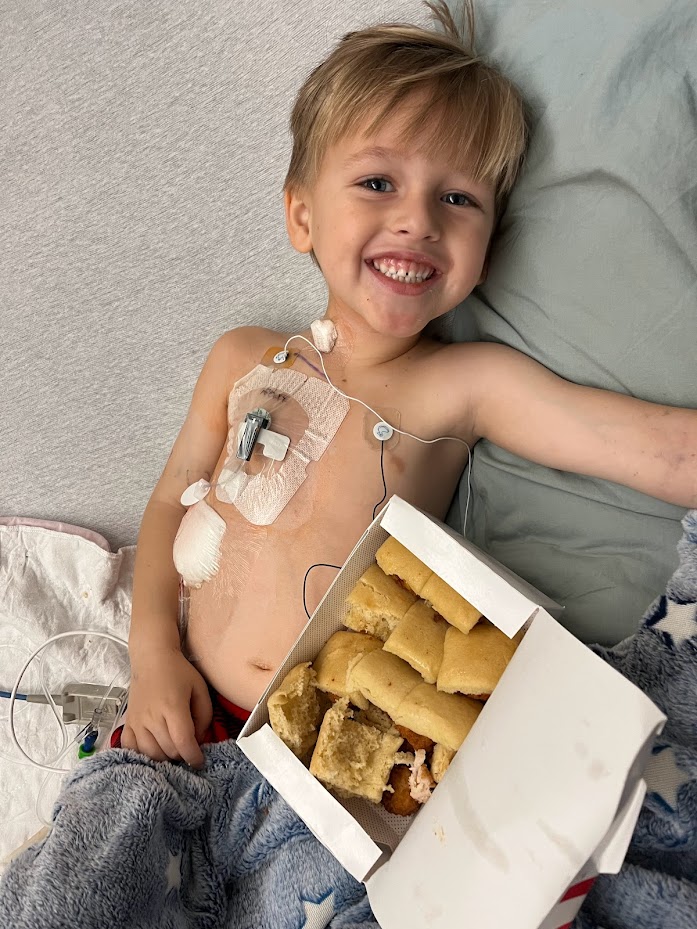 smiling young white boy laying in bed with a port in with a box of rolls