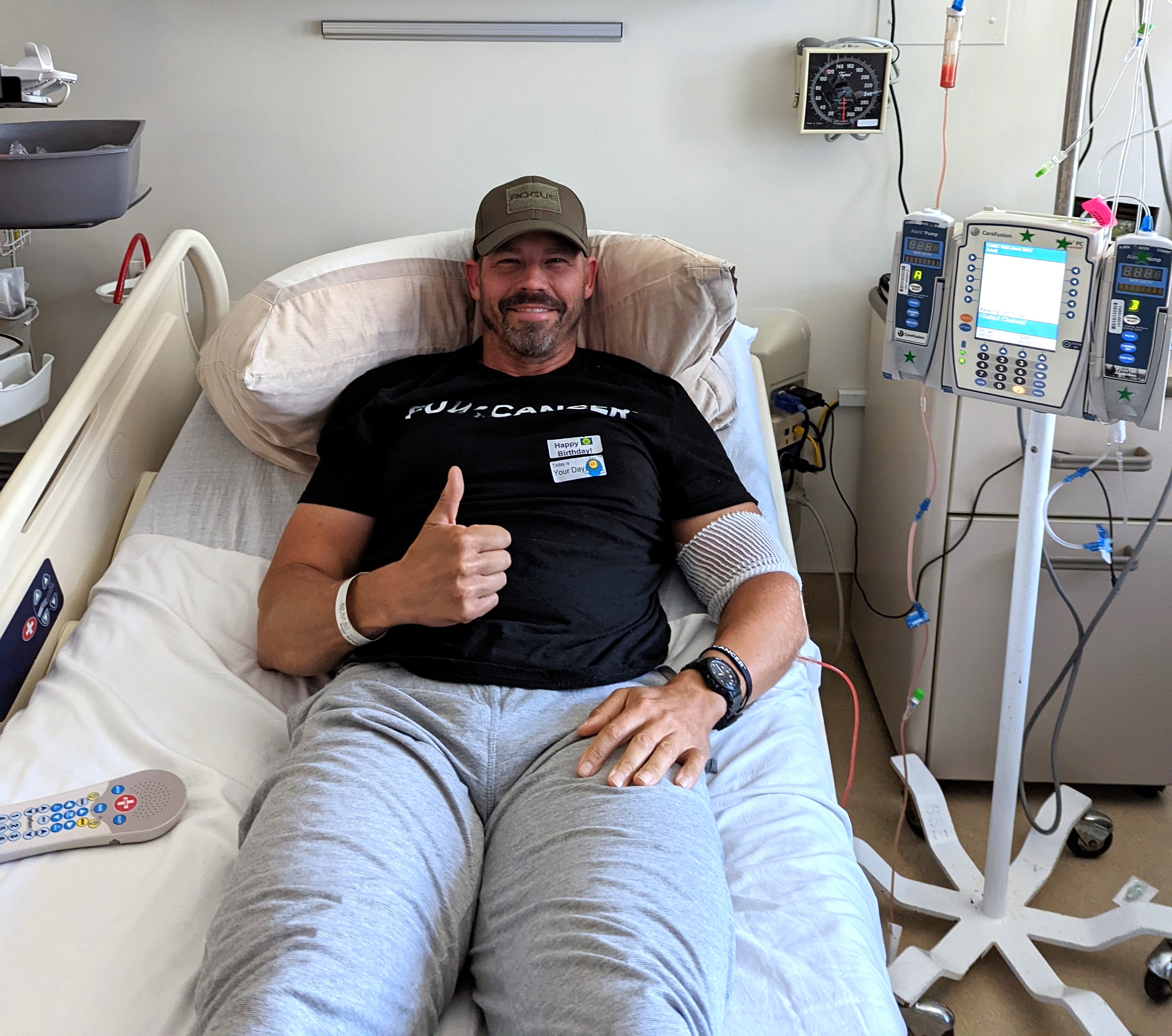 middle aged white man in a ball cap with a scruffy beard and mustached wearing a black t-shirt lying in a hospital bed giving a thumbs up