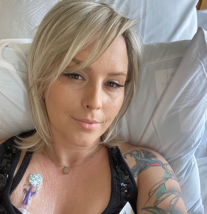 young woman with blond hair and long eyelashes lying in hospital bed wearing a black sleeveless top showing a port and a tattoo