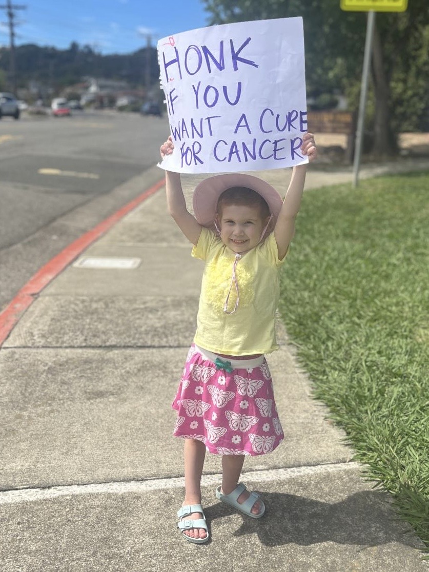 little white girl in a yellow shirt and pink skit with butterflies on it wearing a hat and holding up a sign that says honk if you want a cure for cancer