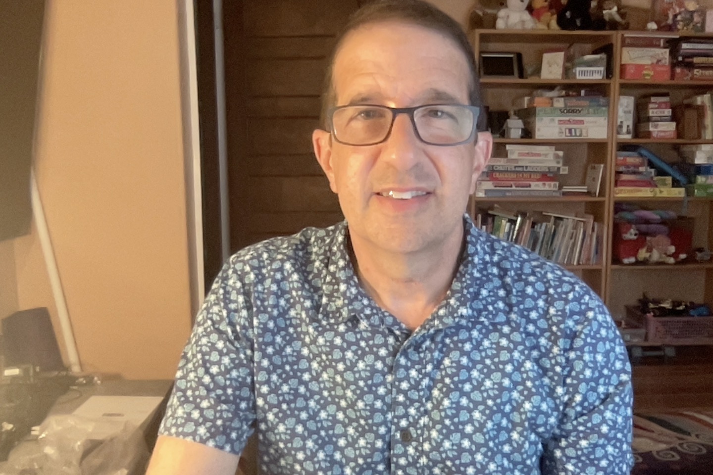 middle aged white man wearing glasses and a blue print shirt sitting in front of a bookcase