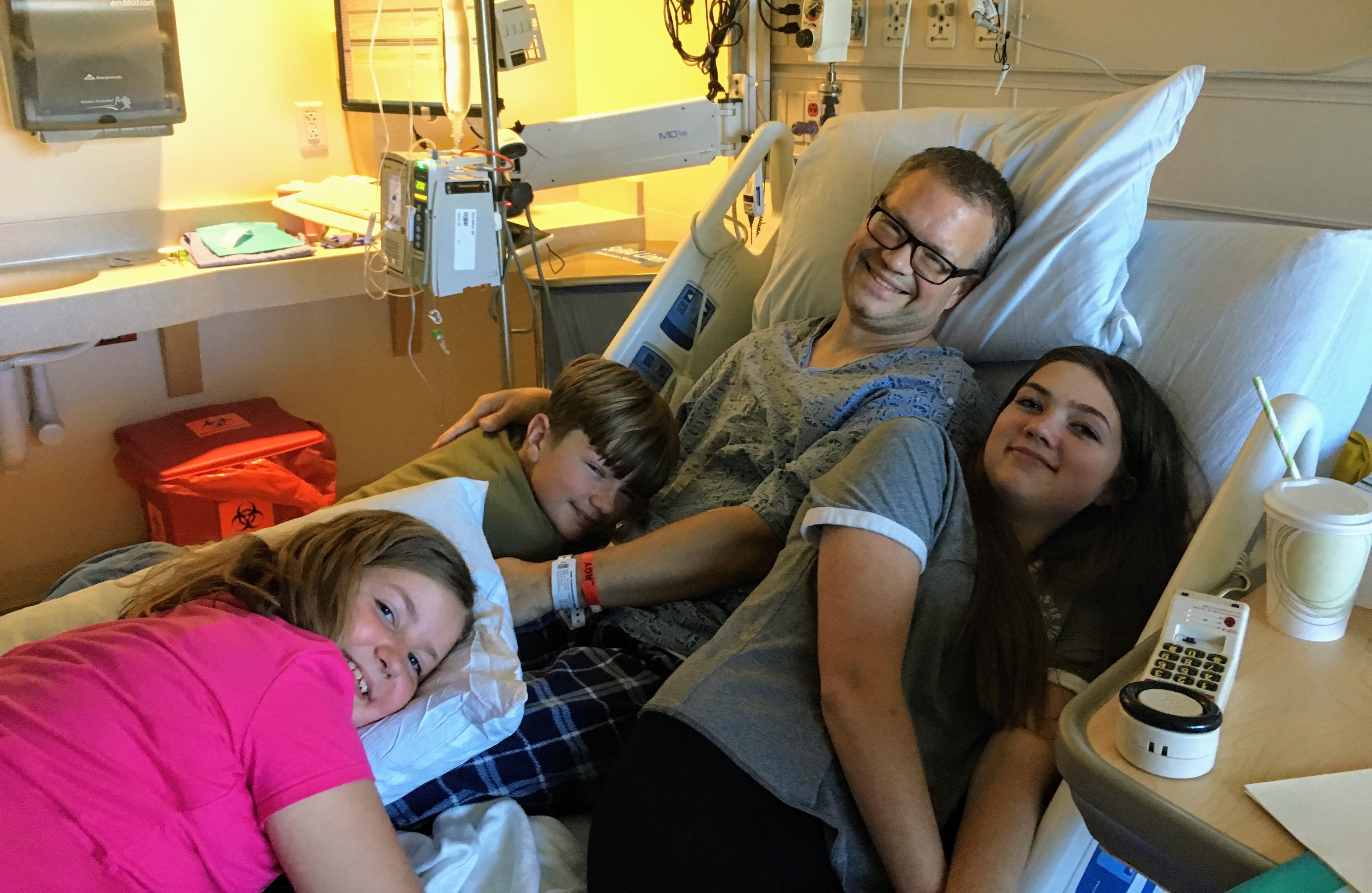 middle aged white man wearing glasses and a hospital gown sitting on a hospital bed with his son and two daughers