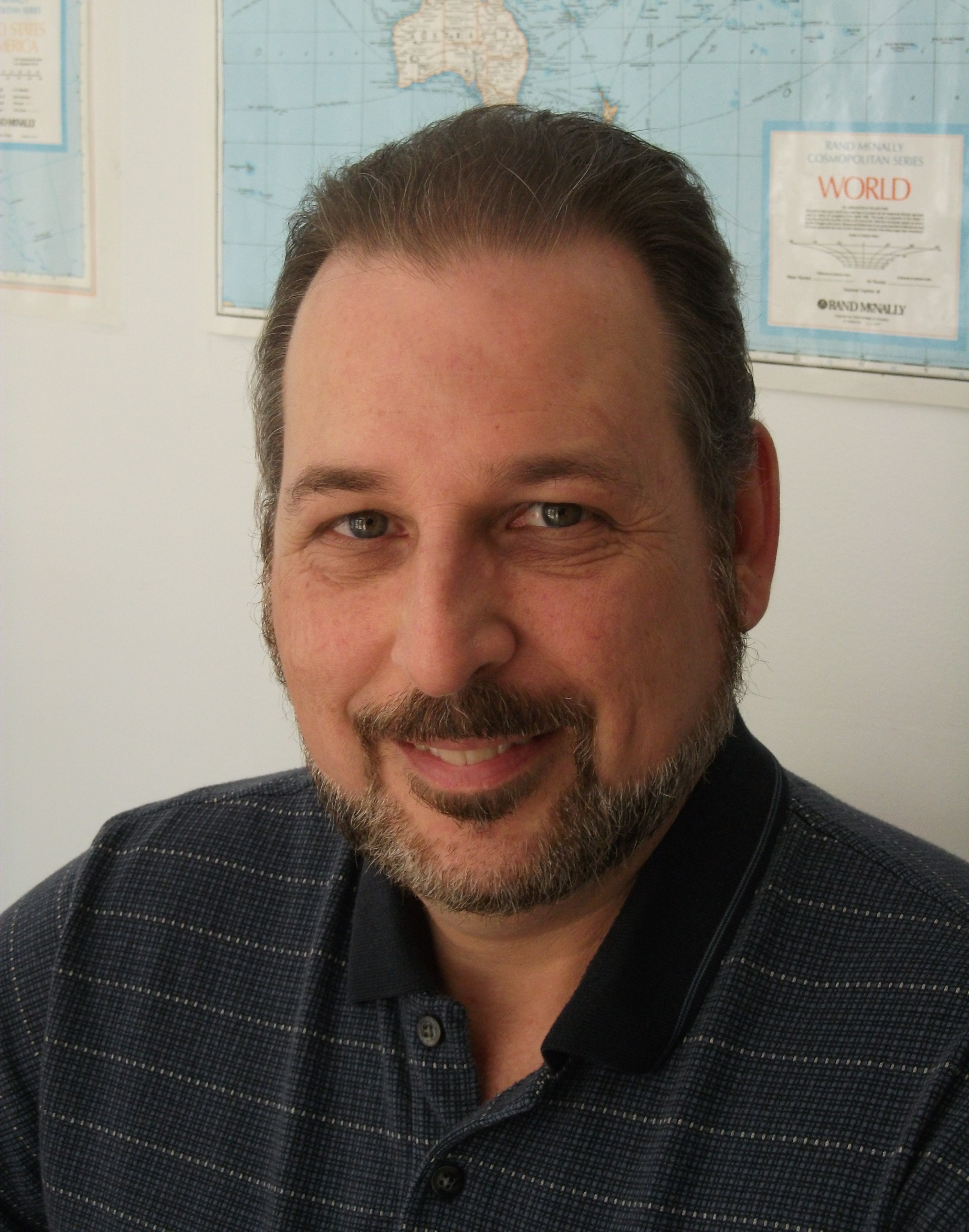 middle aged white man with a beard and mustache wearing a navy blue shirt sitting in front of a world map