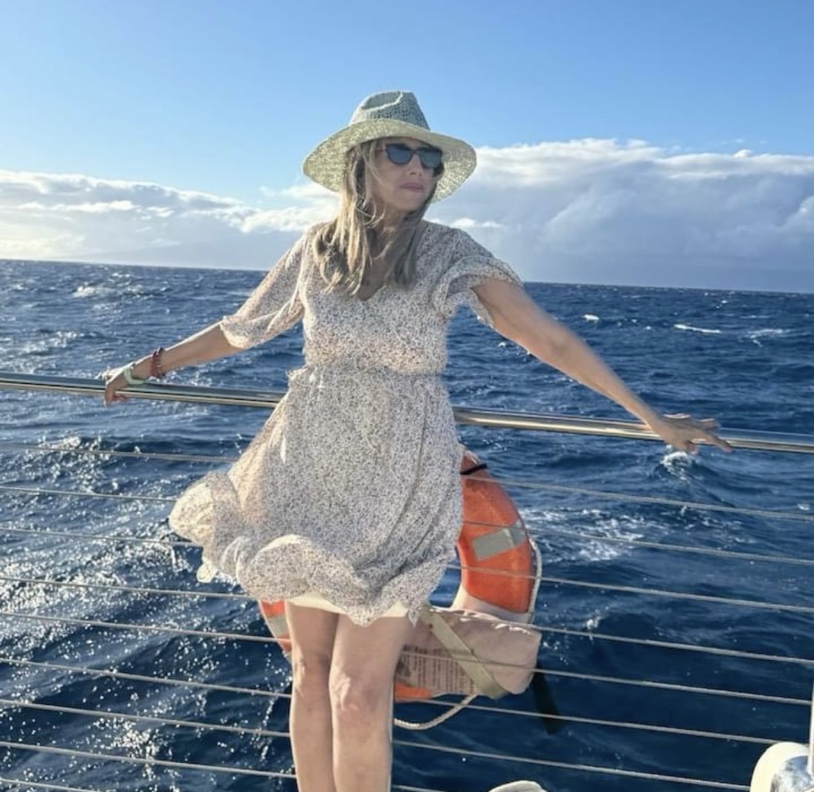 middle aged white woman wearing a straw hat sunglasses and a floral dress leaning on a boat's railing