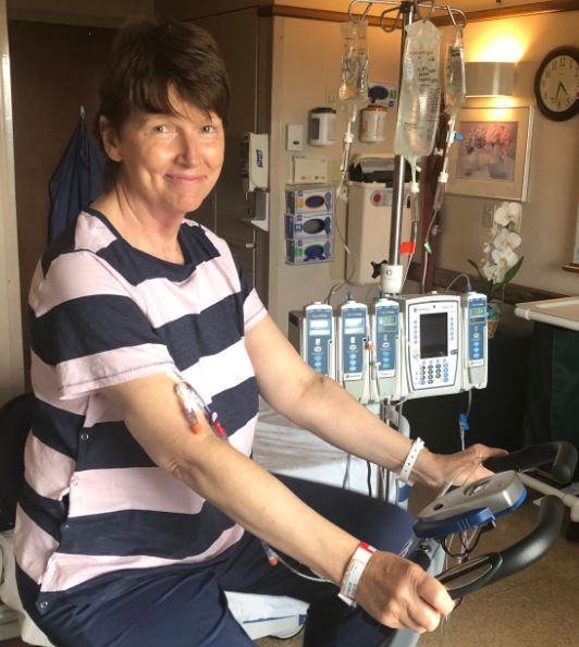 middle aged white woman with short brown hair wearing a navy and pink striped blouse and blue pants riding a stationary bike in a hospital room