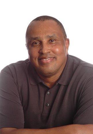 older black man smiling wearing a burgundy colored shirt with his arms crossed