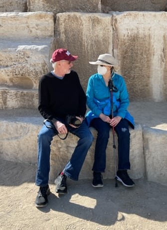 older white couple sitting in front of stones man wearing burgundy ball cap and black shirt and woman wearing blue shirt face maske and straw hat