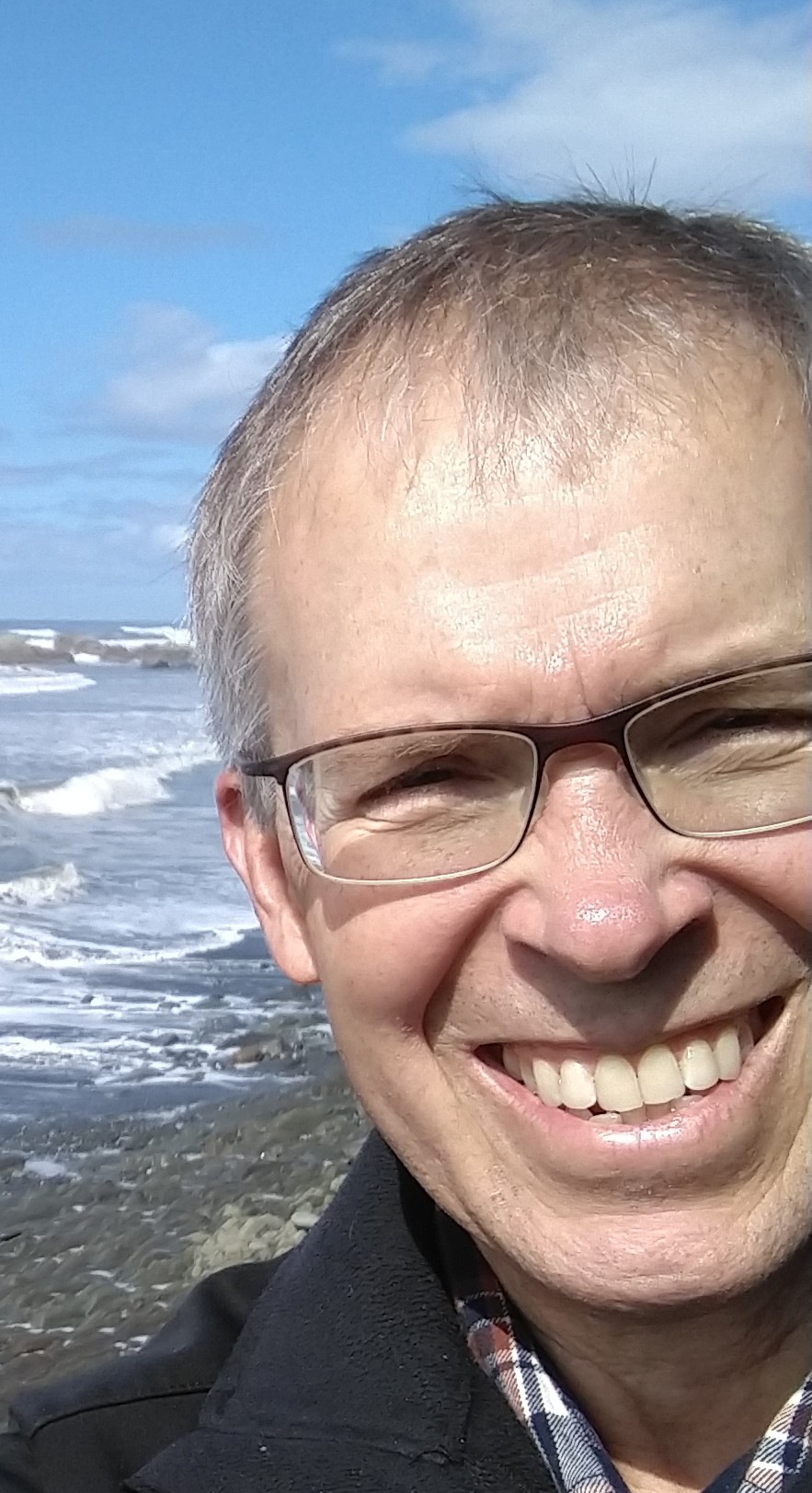 older white man with brown framed glasses plaid shirt and black jacket and big smile standing in front of the ocean