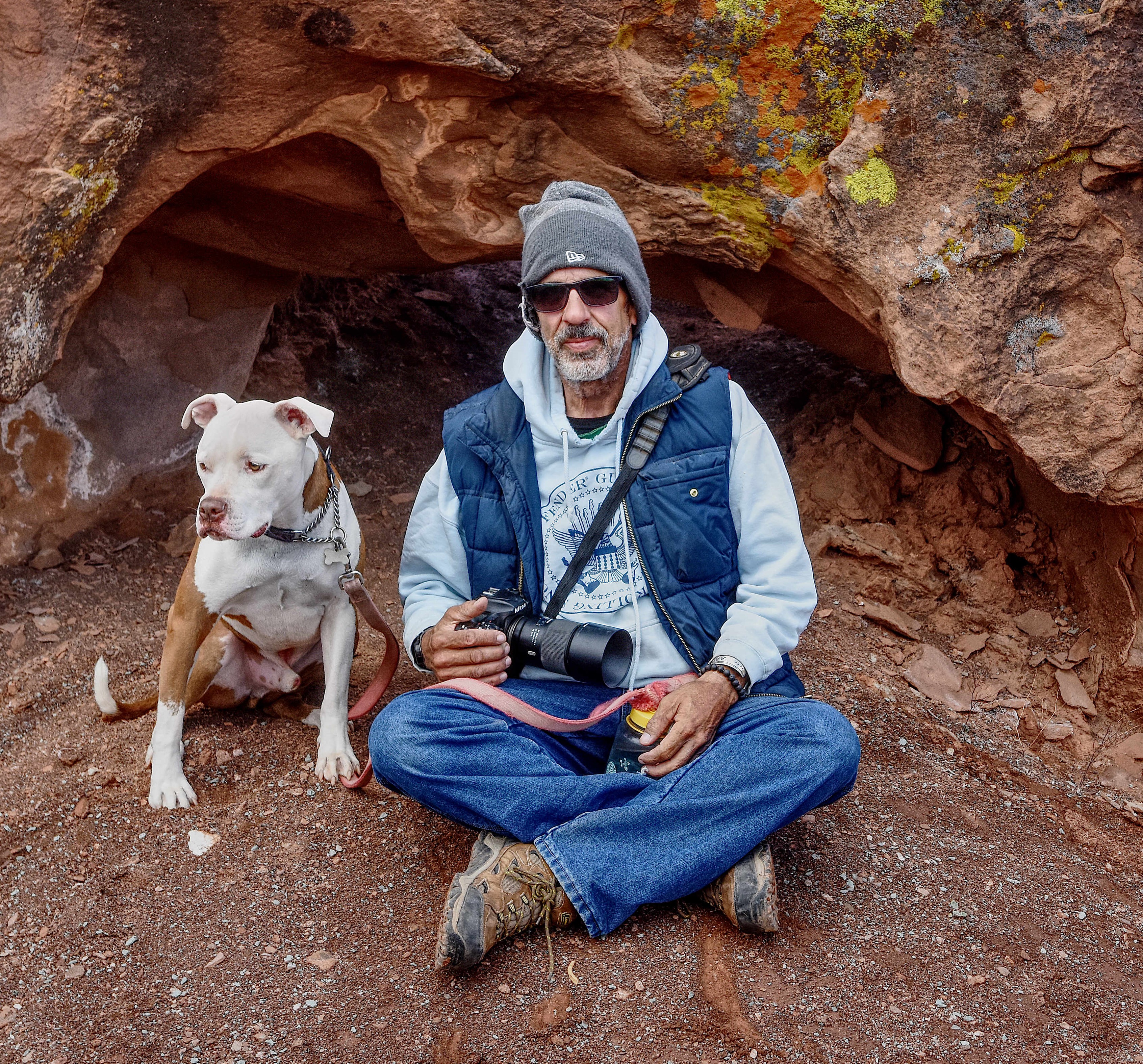 older white man with beard and mustache wearing a knit cap and puffy blue vest holding a camera sitting by his dog