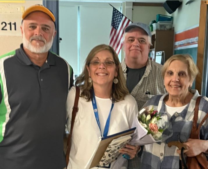 two older men wearing ball caps with a woman wearing glasses and dangly earrings holding a bouquet of flowers and their mom on the right and an American flag in the background