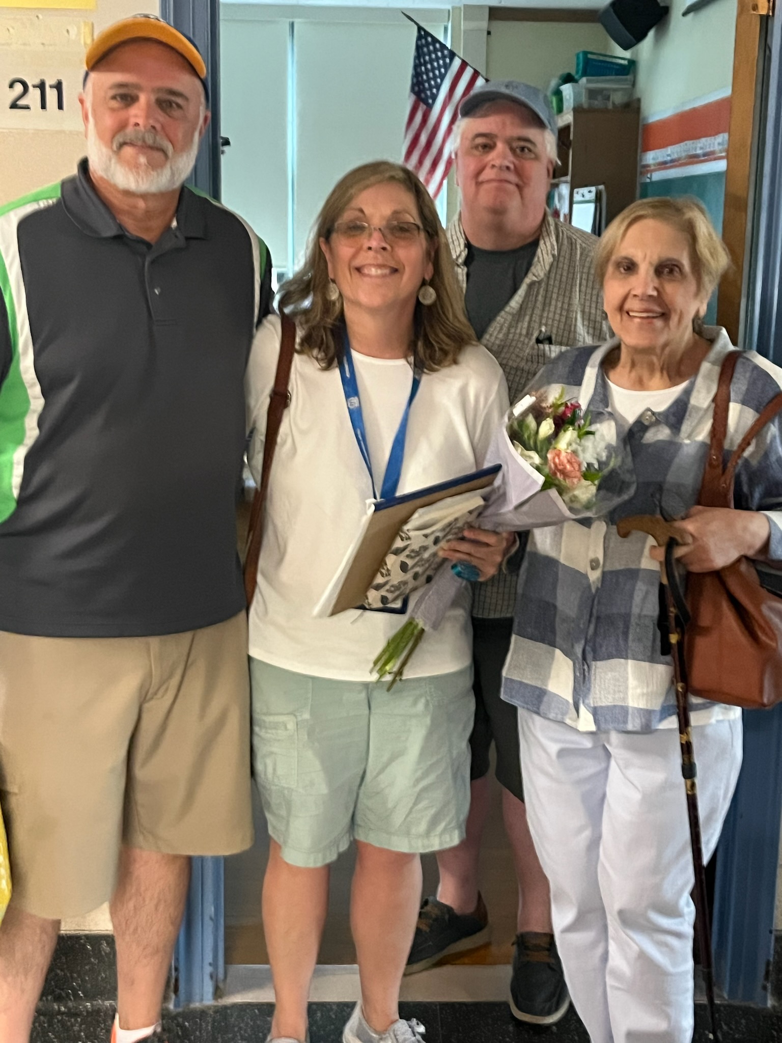 two older men wearing ball caps with a woman wearing glasses and dangly earrings holding a bouquet of flowers and their mom on the right and an American flag in the background