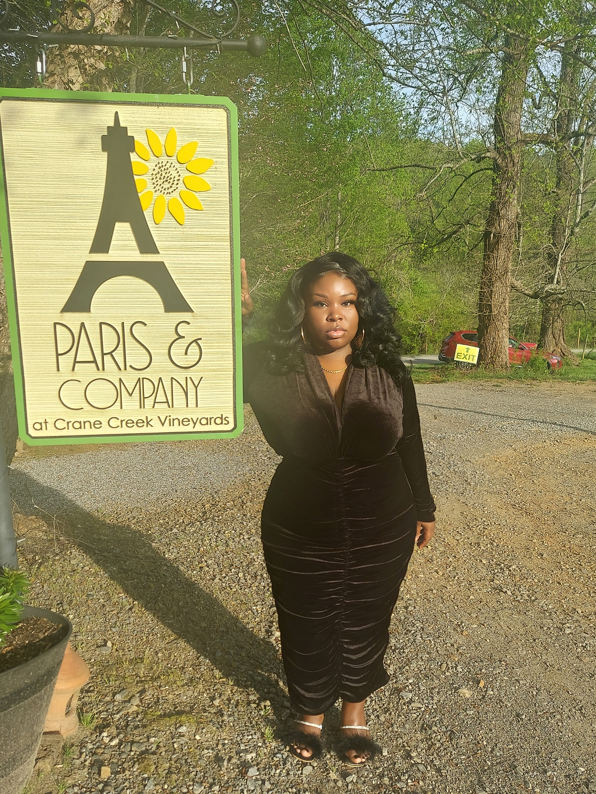 young black woman wearing a black dress with gold hoop earrings and necklace standing by a sign that says Paris and Company