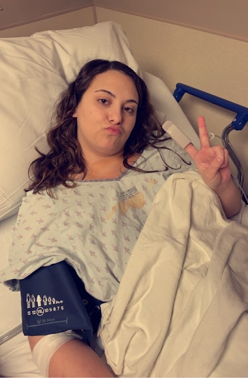 young woman with long brown hair wearing hosptial gown puckering her mouth and giving a peace sign lying in a hospital bed