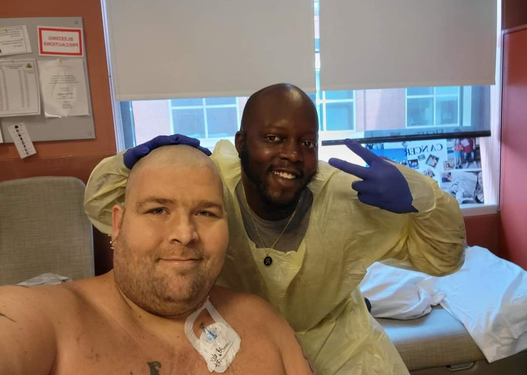 younger white male with shower cap on scruffy beard and no shirt sitting in front of black male medical tech giving a peace sign