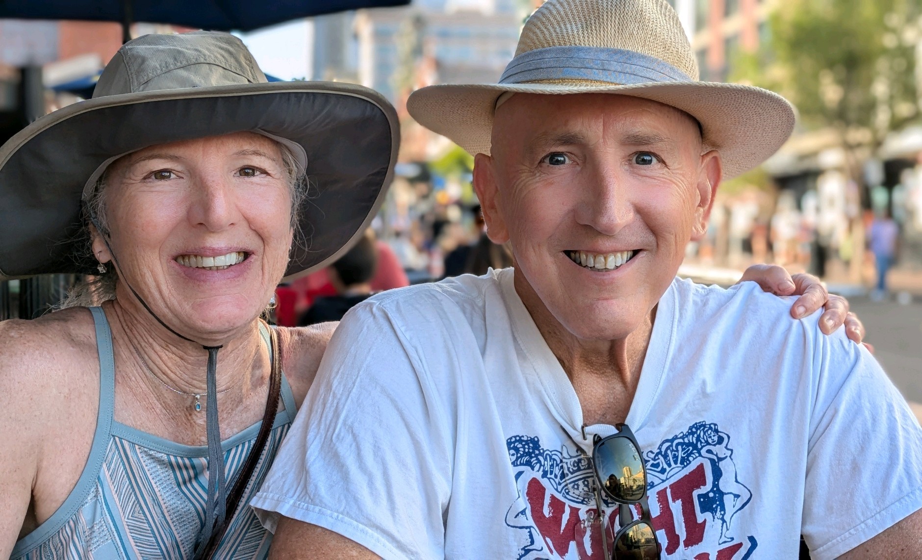 older white couple both wearing hats and smiling man wears white t-shirt with sunglasses hanging from the neck