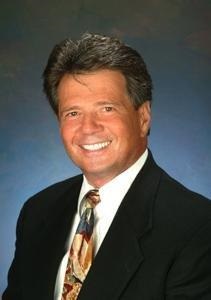 older white man headshot smiling wearing a brown suite white shirt and brown and blue tie