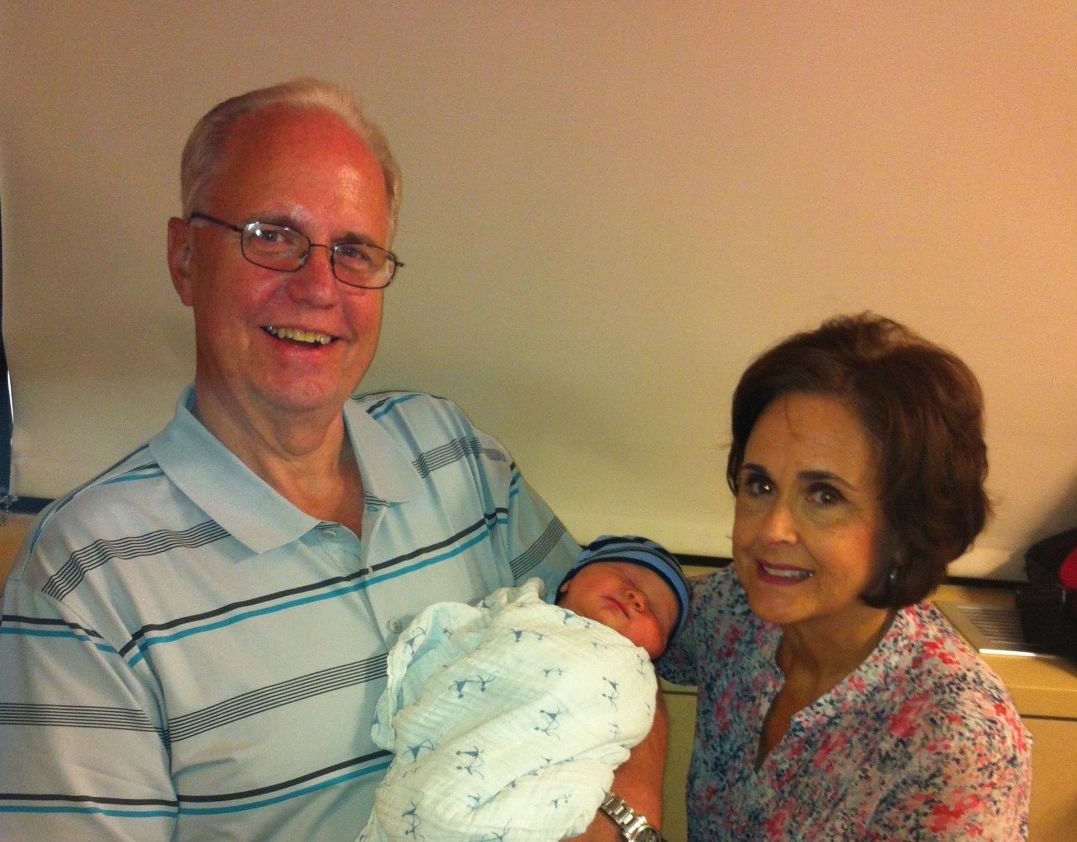 older white man with glasses and blue striped shirt holding a baby with an older woman with dark hair and a floral blouse