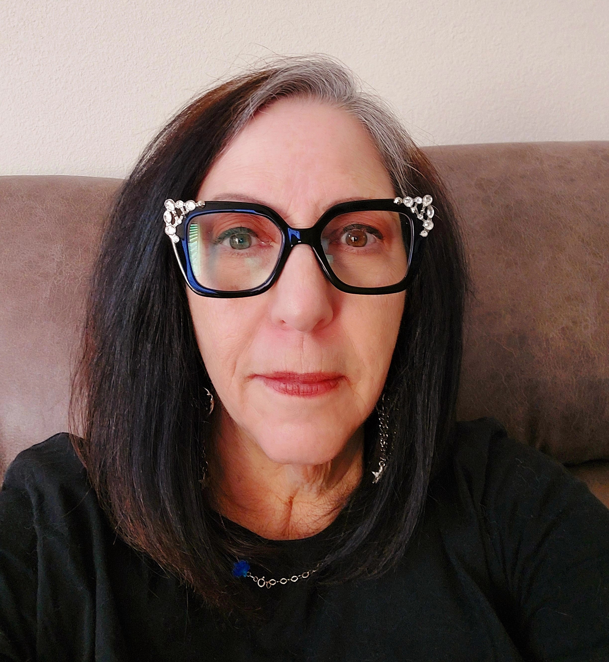 older white woman with dark long hair wearing black chunky glasses with bling on them and a black top