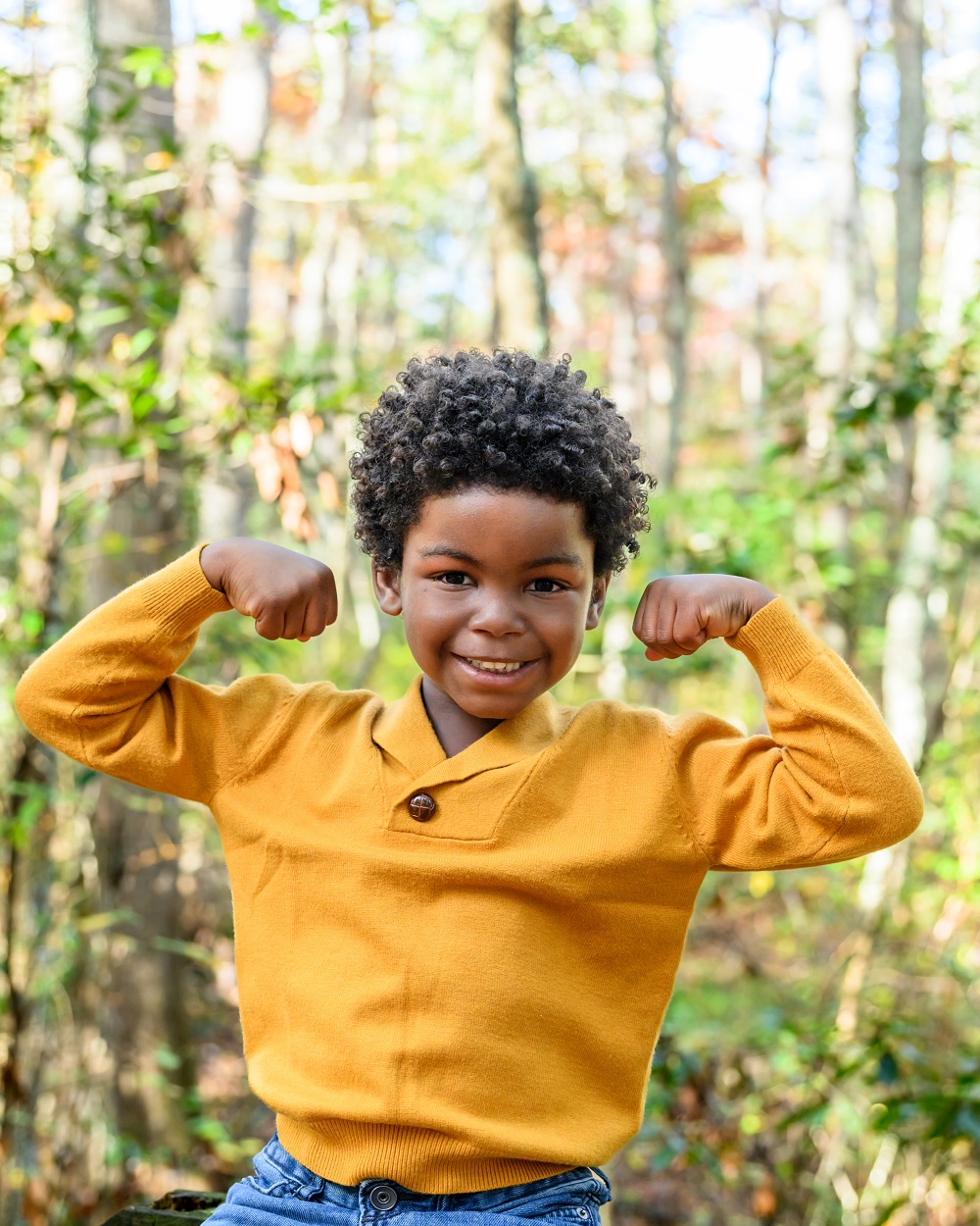 smiling young black boy standing in front of trees wearing a gold sweater and smiling and making muscle man arms