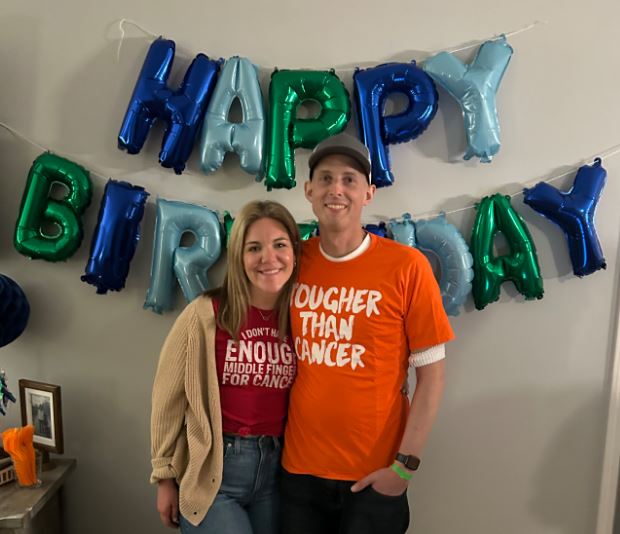 young white couple standing in front of a birthday balloon sign she has blond hair and burgundy t-shirt on he is wearing a ball cap and orange t-shirt