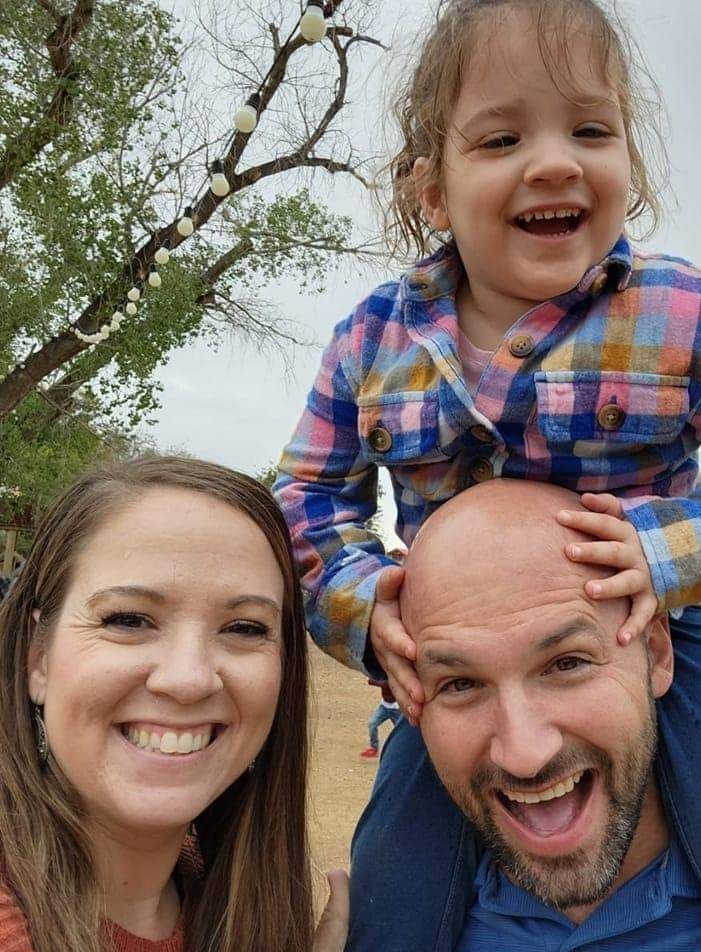 young white couple with their daughter outside little girl has on plaid shirt dad is bald with a mustache and beard mom has long brown hair