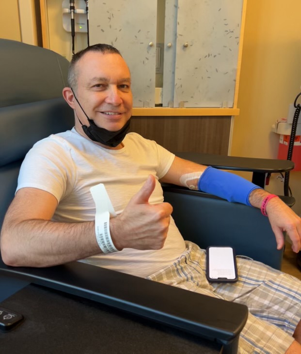 middle age white man with short hair, a black mask pulled down, a blue sleeve on his arm giving a thumbs up wearing white shirt with plaid shorts