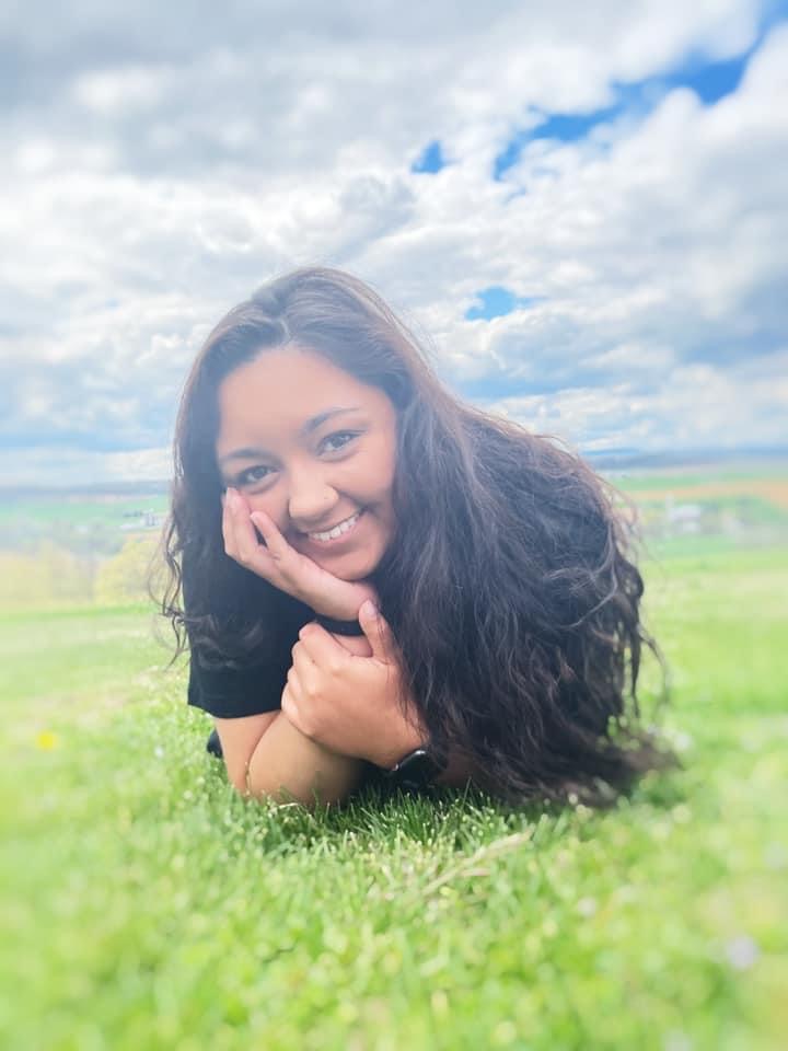 smiling young hispanic girl with long dark hair lying in the grass head propped on hand wearing black shirt and watch