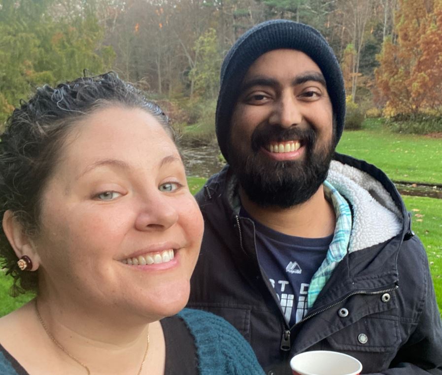 young white woman in blue sweater holding coffee with young black man with beard and mustache wearing a knit cap and blue jacket holding a cup