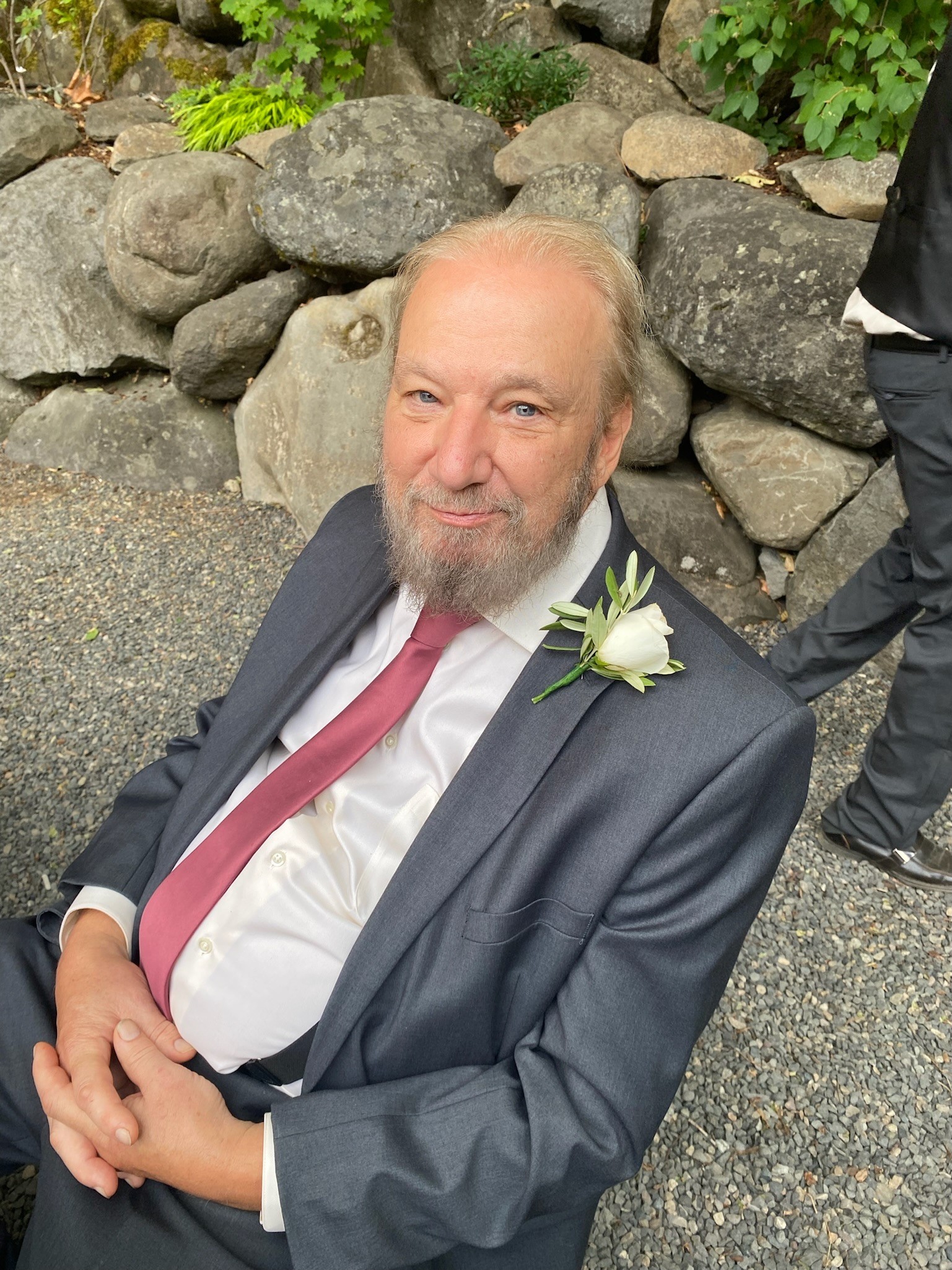 Gary older white man with blue eyes and thinning hair and gray beard and mustache leaning on rocks wearing a blue suite, mauve tie and a rose boutonaire