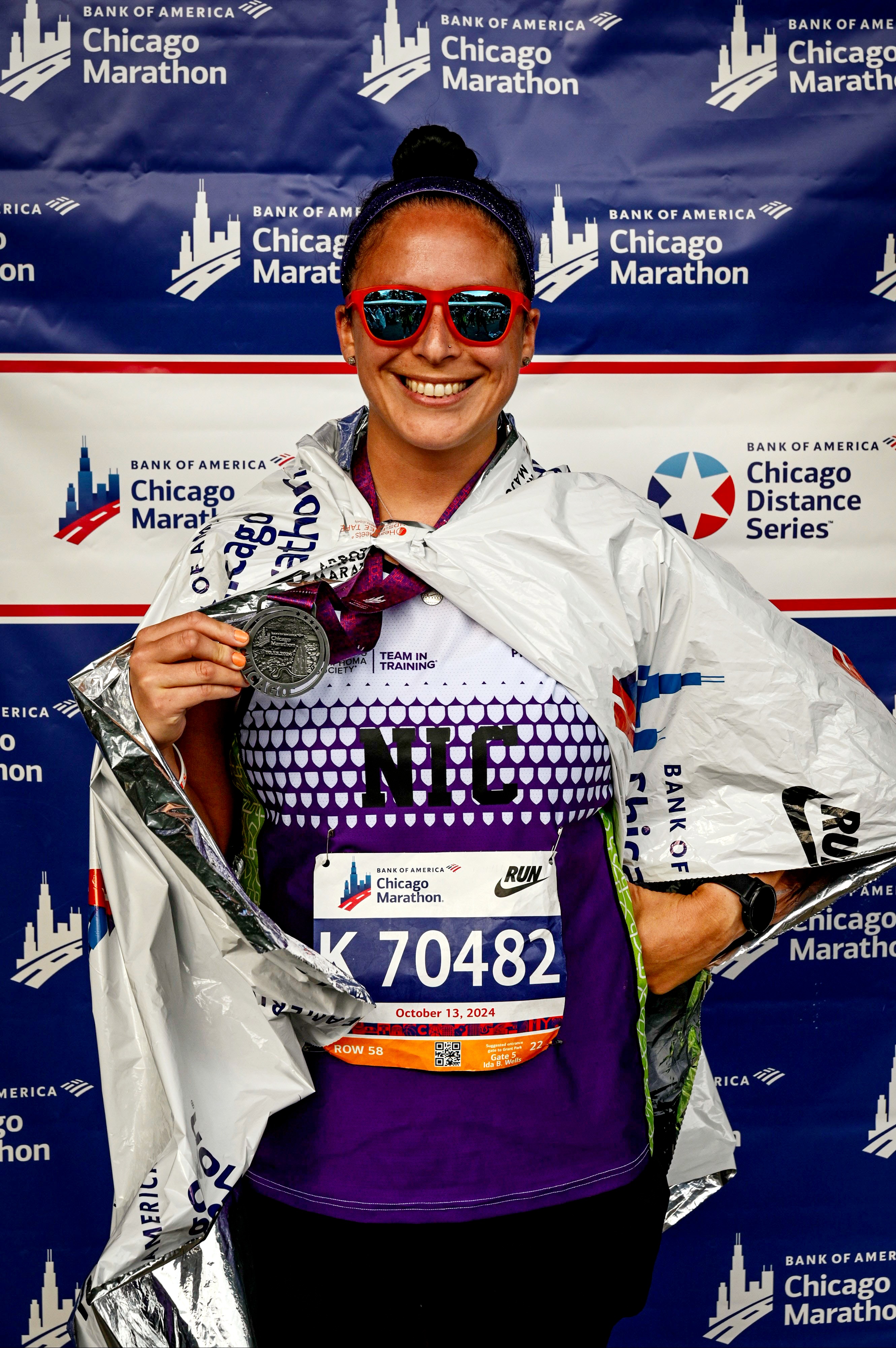 Nicolas smiling middle aged hispanic woman with dark hair pulled up in a bun wearing orangish sunglasses and a Team In Training Shirt holding a medallion in front of a Chicago Marathon banner.jpg